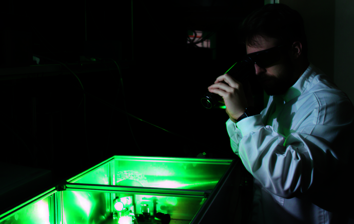 Ein Mann mit Laserschutzbrille und weißer Schutzkleidung blickt durch ein optisches Messgerät auf eine grün leuchtende Box. Ein Mann mit Laserschutzbrille und weißer Schutzkleidung blickt durch ein optisches Messgerät auf eine grün leuchtende Box.