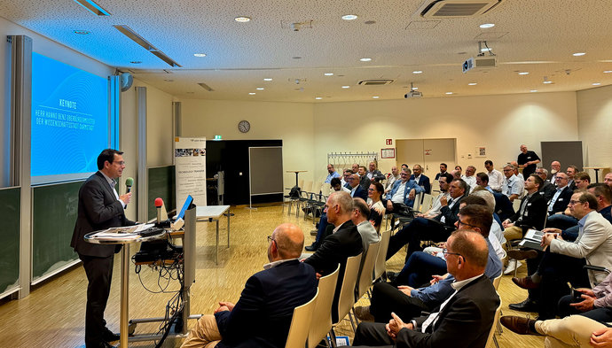 Ein Mann steht vor einer Leinwand an einem Pult. Vor ihm sitzt eine Gruppe Personen in einem Vortragsraum. Ein Mann steht vor einer Leinwand an einem Pult. Vor ihm sitzt eine Gruppe Personen in einem Vortragsraum.