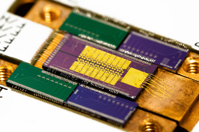 Auf einem kupferfarbenen Board ist mittig ein rechteckiger Mikrochip mit goldenen Verdrahtungen zu sehen. Er ist umgeben von vier kleineren grünen und blauen Chips. Auf einem kupferfarbenen Board ist mittig ein rechteckiger Mikrochip mit goldenen Verdrahtungen zu sehen. Er ist umgeben von vier kleineren grünen und blauen Chips.